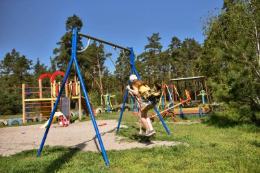 İki sarışın liseli kız salıncakta sallanıyor bir çocuk parkında çam ormanının yakınında
