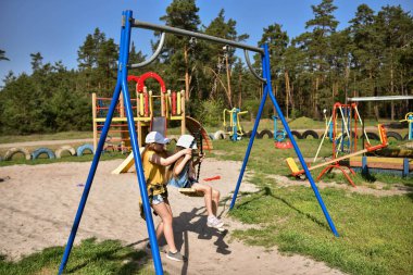 İki sarışın liseli kız salıncakta sallanıyor bir çocuk parkında çam ormanının yakınında