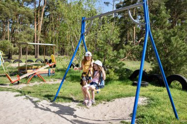 İki sarışın liseli kız salıncakta sallanıyor bir çocuk parkında çam ormanının yakınında