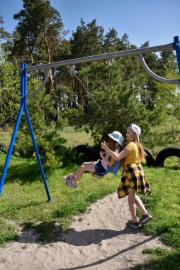 İki sarışın liseli kız salıncakta sallanıyor bir çocuk parkında çam ormanının yakınında