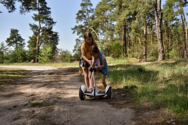 İki liseli kız, çam ormanında bir patikada scooter kullanıyor.