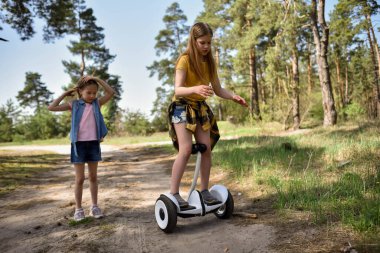 İki liseli kız, çam ormanında bir patikada scooter kullanıyor.