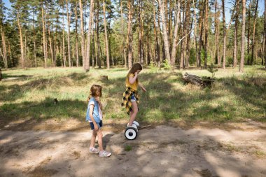 İki liseli kız, çam ormanında bir patikada scooter kullanıyor.
