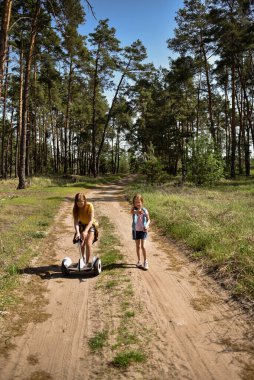 İki liseli kız, çam ormanında bir patikada scooter kullanıyor.