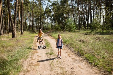 İki liseli kız, çam ormanında bir patikada scooter kullanıyor.