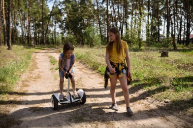 İki liseli kız, çam ormanında bir patikada scooter kullanıyor.