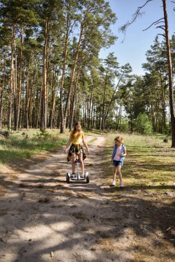 İki liseli kız, çam ormanında bir patikada scooter kullanıyor.
