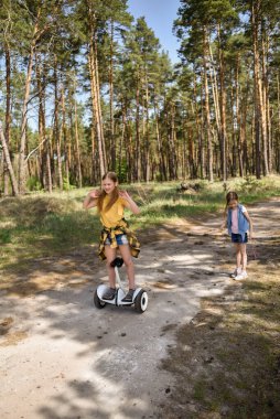 İki liseli kız, çam ormanında bir patikada scooter kullanıyor.