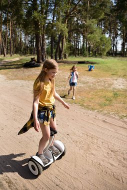 İki liseli kız, çam ormanında bir patikada scooter kullanıyor.
