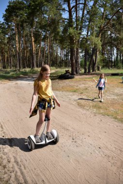 İki liseli kız, çam ormanında bir patikada scooter kullanıyor.