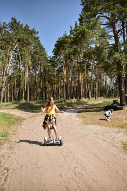 İki liseli kız, çam ormanında bir patikada scooter kullanıyor.
