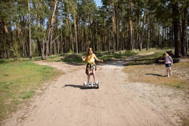 İki liseli kız, çam ormanında bir patikada scooter kullanıyor.