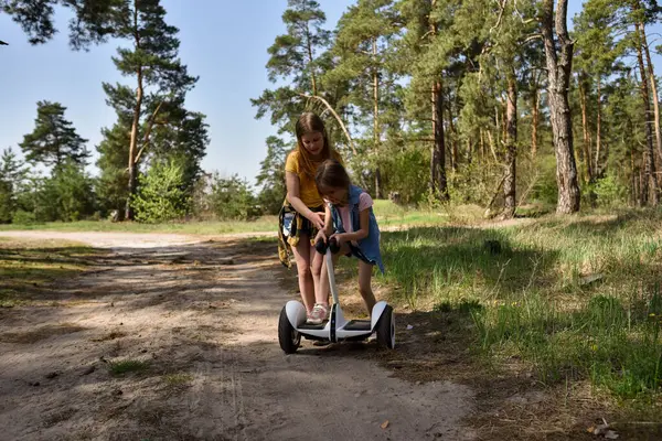 İki liseli kız, çam ormanında bir patikada scooter kullanıyor.