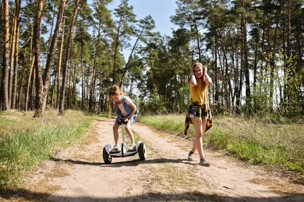 İki liseli kız, çam ormanında bir patikada scooter kullanıyor.