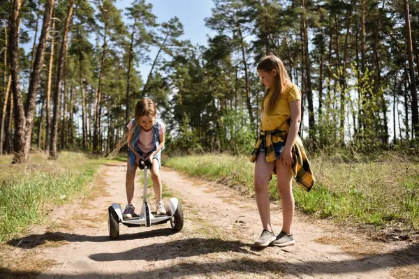 İki liseli kız, çam ormanında bir patikada scooter kullanıyor.