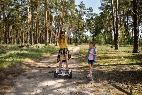İki liseli kız, çam ormanında bir patikada scooter kullanıyor.