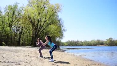Spor takım elbiseli iki genç kız şehir parkında nehrin kenarında fiziksel egzersiz yapıyorlar.
