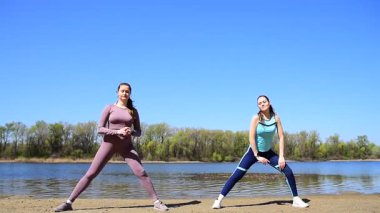 Spor takım elbiseli iki genç kız şehir parkında nehrin kenarında fiziksel egzersiz yapıyorlar.