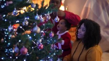 Yandan bakınca gülümseyen Afrikalı Amerikalı genç bir kadın Noel ağacını Noel arifesinde erkek ve küçük bir çocukla süslüyor. Mutlu ve rahat bir aile evde yeni yıl hazırlıklarının tadını çıkarıyor.