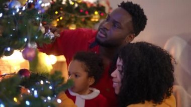 Mutlu Afro-Amerikan genç ailesi Noel ağacını evde süslüyor. Gülümseyen, rahat kadın ve küçük çocuğun yeni yıl ağacı dekorasyonundan bahseden portresi.