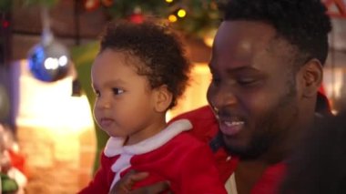 Gülümseyen neşeli çocuk Noel ağacında yeni yıl oyuncağıyla oynarken gülümseyen genç adam konuşuyor. Afrika kökenli Amerikalı baba ve oğlunun yakın plan portresi.
