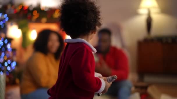 可爱的男孩看着镜头 转身走向坐在客厅壁炉边的笑着的年轻父母 迷人的非洲裔美国幼儿与父母欢度新年的肖像 — 图库视频影像
