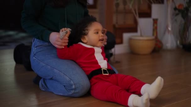 サンタクロースの衣装でかわいいアフリカ系アメリカ人の男の子と遊ぶ認識できない若い白人女性 正の叔母楽しむクリスマスレジャーで甥と屋内でリビングルーム — ストック動画