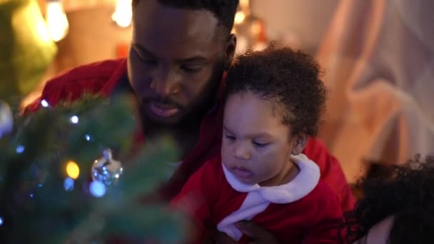 クローズアップは 自宅で両親とクリスマスツリーを飾る小さな男の子のキュリオスを集中 高角度ビューポルノのアフリカ系アメリカ人の息子楽しんで新年前夜とともに父接吻子供の頬 — ストック動画