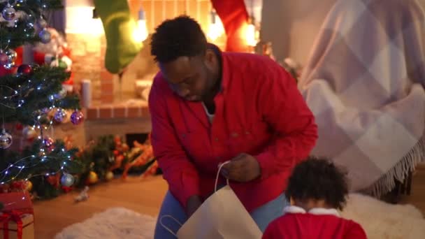 Ung Afroamerikansk Man Lägger Julklappar Nyårsträdet Mysigt Vardagsrum Som Nyfikna — Stockvideo
