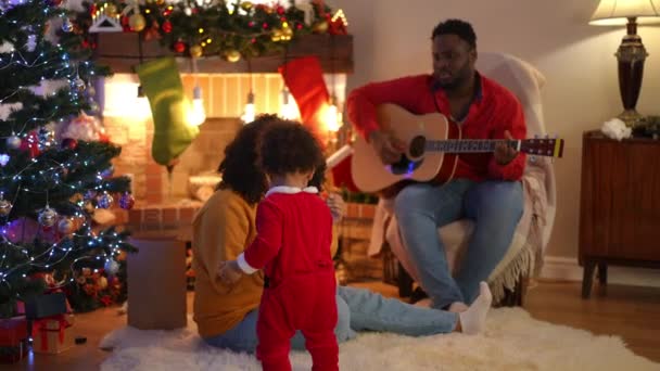 広いショット若いアフリカ系アメリカ人の母親は暖炉でバックグラウンドでギターを弾く父親の夫として幼児の息子と遊んでいます 一緒に家でクリスマスを祝う幸せな自信のある家族 — ストック動画