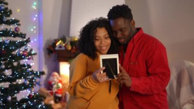 Noel 'de evde dikilen Afro-Amerikalı kadın ve erkeğin fotoğraf çerçevesine hayranlıkla sarılırken çekilmiş orta boy bir fotoğraf. Mutlu genç karı koca sohbetleri gülümseyerek yeni yıl arifesinin tadını çıkarıyor.