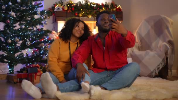 愛するアフリカ系アメリカ人の若いカップルは クリスマスの前夜に暖炉に座ってスマートフォンに自撮りを取ります 家族を誇るリラックスしたポジティブな男と女の広いショットのポートレート新年ソーシャルメディアで — ストック動画
