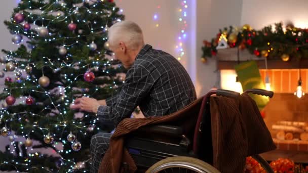除夕之夜 一位头发灰白的老人坐在轮椅上 在客厅里装饰着新年的圣诞树 白人退休人员在室内享受圣诞准备的侧面视角 生活方式和欢乐 — 图库视频影像