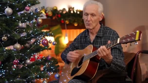 除夕之夜 坐在轮椅上专注于音乐的灰发音乐家在家里弹奏吉他 集中的白人老人在客厅里享受圣诞情趣的画像 — 图库视频影像