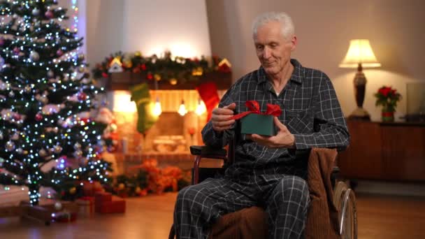 坐在轮椅上心满意足的老人的画像 客厅里坐着圣诞礼物 笑着思考 佳节前夕 心情愉快的白种人退休人员带着新年礼物回家 — 图库视频影像