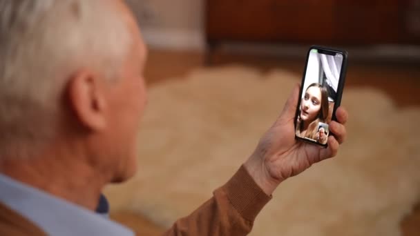 シニア男性の手と喜びの十代の女の子の話でビデオチャットでスマートフォン モバイルアプリケーションで孫娘とオンラインで白人の祖父メッセージングの肩の上に撮影 — ストック動画