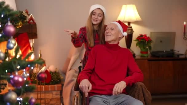 明るい10代の少女が車椅子で高齢者と話をし リビングルームでクリスマスツリーを賞賛しました 白人の孫娘と祖父の家で一緒に新年を楽しむ肖像画 — ストック動画