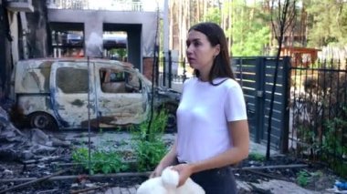 Yan görünüşü muhteşem, ince, Ukraynalı kadın. Ön bahçede oyuncak ayıyla dikilip bombayla yıkılmış eve bakıyor. Çaresiz genç bir bayanın portresi savaşta kaybolan mallar.