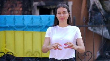 Yıkılmış evdeki çitlerin üzerinde Ukrayna bayrağının arka planında kalp şeklinde jestler yapan kendine güvenen güzel bir kadın. Dışarıda poz veren cılız ve güzel bir bayanın portresi. Destek için teşekkür ediyor.