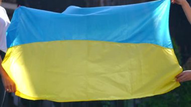 Yakın plan Ukrayna bayrağı beyaz kadınlarda ağır çekimde pohpohlanıyor. Bombalanmış şehrin güneşli havasında ellerinde sarı ve mavi ulusal bayrak tutan tanınmayan iki genç kadın.