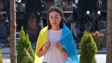 Ulusal bayrağa sarılmış kameraya gülümseyen cesur, güzel Ukraynalı genç kadın. Mahvolmuş şehir caddesinde yanan evin arka planında poz veren muhteşem cesur kadının portresi.