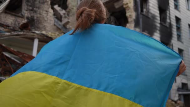 破壊された民家を屋外で見て肩の上にウクライナのフラグを持つ女性の後ろのビューを傾ける キエフの住宅ビルを破壊爆撃で若い女性のバックビューウクライナ — ストック動画