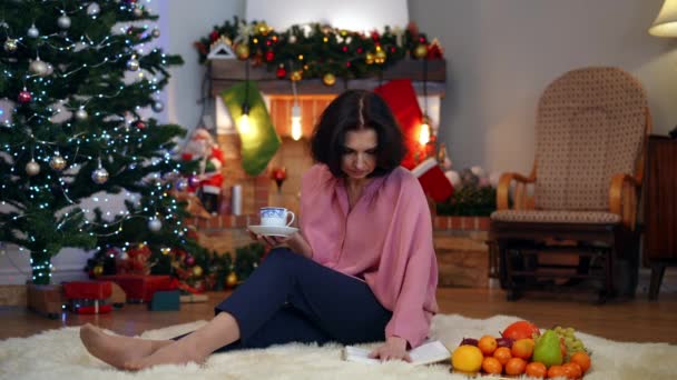 在圣诞前夕 一个人坐在家里 一边看书 一边喝着茶杯 一边望着远方 那是个大快朵颐的女人 描写悲伤的白人孤独寂寞的女士在家里过年的形象 孤独和悲伤 — 图库视频影像