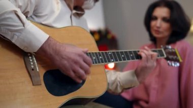 Erkek eli çalan yakın çekim gitar ve arka planda müzik dinleyen bulanık kadın. Kafkasyalı tanınmayan koca, Noel arifesinde evde karısına serenat yapıyor.