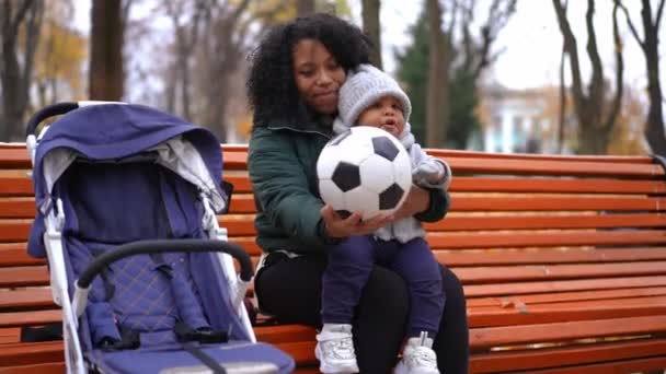 幼い息子と一緒に秋の公園のベンチに座ってサッカーボールと話している若い幸せな母親 リラックスしたアフリカ系アメリカ人の女性と男の子の肖像屋外でレジャーを楽しむ — ストック動画