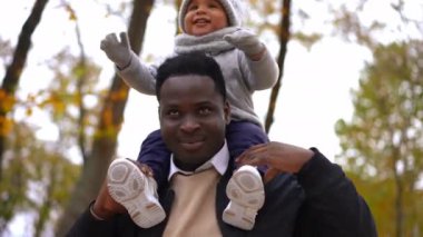 Omuzlarında sevimli bir çocukla kameraya bakıp gülümseyen mutlu Afro-Amerikan gencin portresi. Kendine güvenen baba, sonbahar parkında oğluyla birlikte poz veriyor.