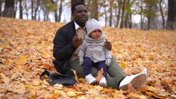 年轻的非裔美国父亲一边看着他一边说着话 一边和刚出生的小孩玩着黄色落叶游戏 在公园里与男孩坐在一起享受周末闲暇的松懈男子 — 图库视频影像