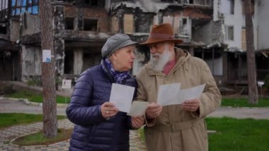 Üst düzey üzgün adam ve kadın arka planda yanmış evlerin fotoğraflarıyla dışarıda konuşuyorlar. Bombalanmış Ukrayna şehrindeki anılarını hatırlatan yaşlı bir çiftin portresi. Yavaş çekim