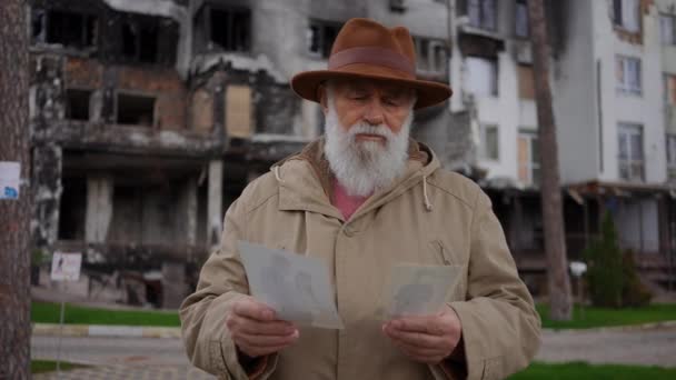 照片中的老年人绝望地看着站在乌克兰城市被轰炸建筑背景下的旧照片 在荒废的城市里沮丧沮丧的退休人员 在户外想别的事 — 图库视频影像