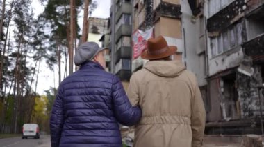 Arkaya bakan yaşlı bir adam ve bir kadın el ele yürüyorlar ve bombalanan evlere bakıyorlar. Yaşlı bir çiftin Ukrayna 'nın harap olmuş şehrinde ağır çekimde gezinirken çekilmiş görüntüleri.